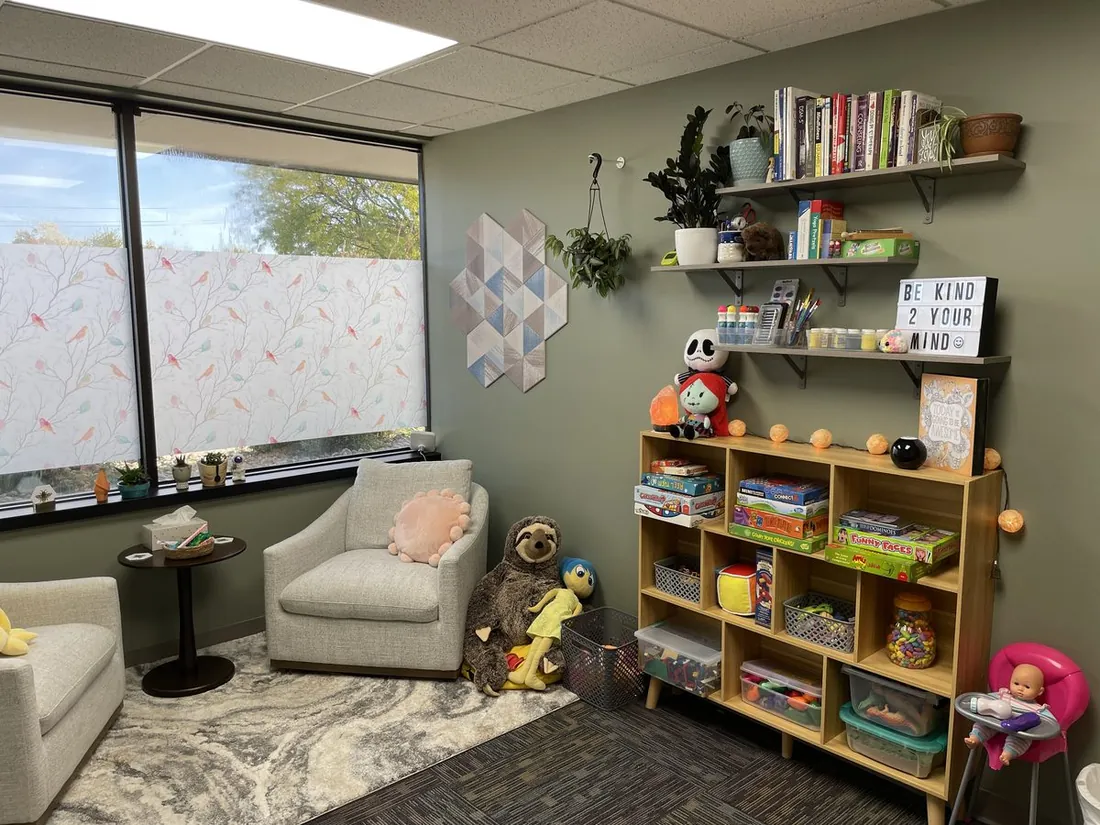 Shelves of clinical books, craft supplies, and organized toys beside cozy seating, with a 'Be Kind 2 Your Mind' sign